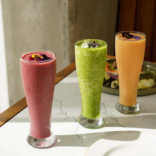 Three colorful smoothies in tall glasses on a marble table, pink, green, and orange, garnished with fruit, sunny casual vibe.