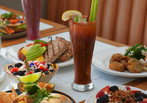 Brunch spread with smoothies, a fresh fruit salad, and plates of eggs, potatoes, and grilled meat on a table.