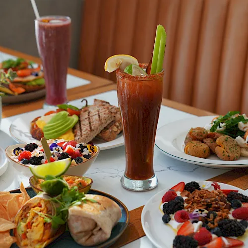 Brunch spread with smoothies, a fresh fruit salad, and plates of eggs, potatoes, and grilled meat on a table.