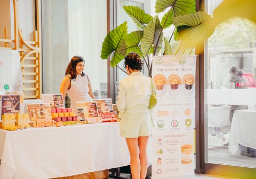 A woman stands at a booth with skincare products, chatting with a vendor inside a bright indoor market setup.