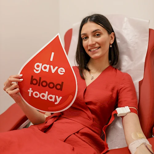 A woman in a red outfit sits in a red chair, holding a sign that says “I gave blood today.”