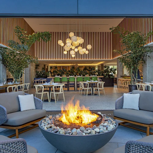 An outdoor seating area with a fire pit, lounge chairs, and a bright indoor dining space, plants, and warm lighting—cozy and inviting.